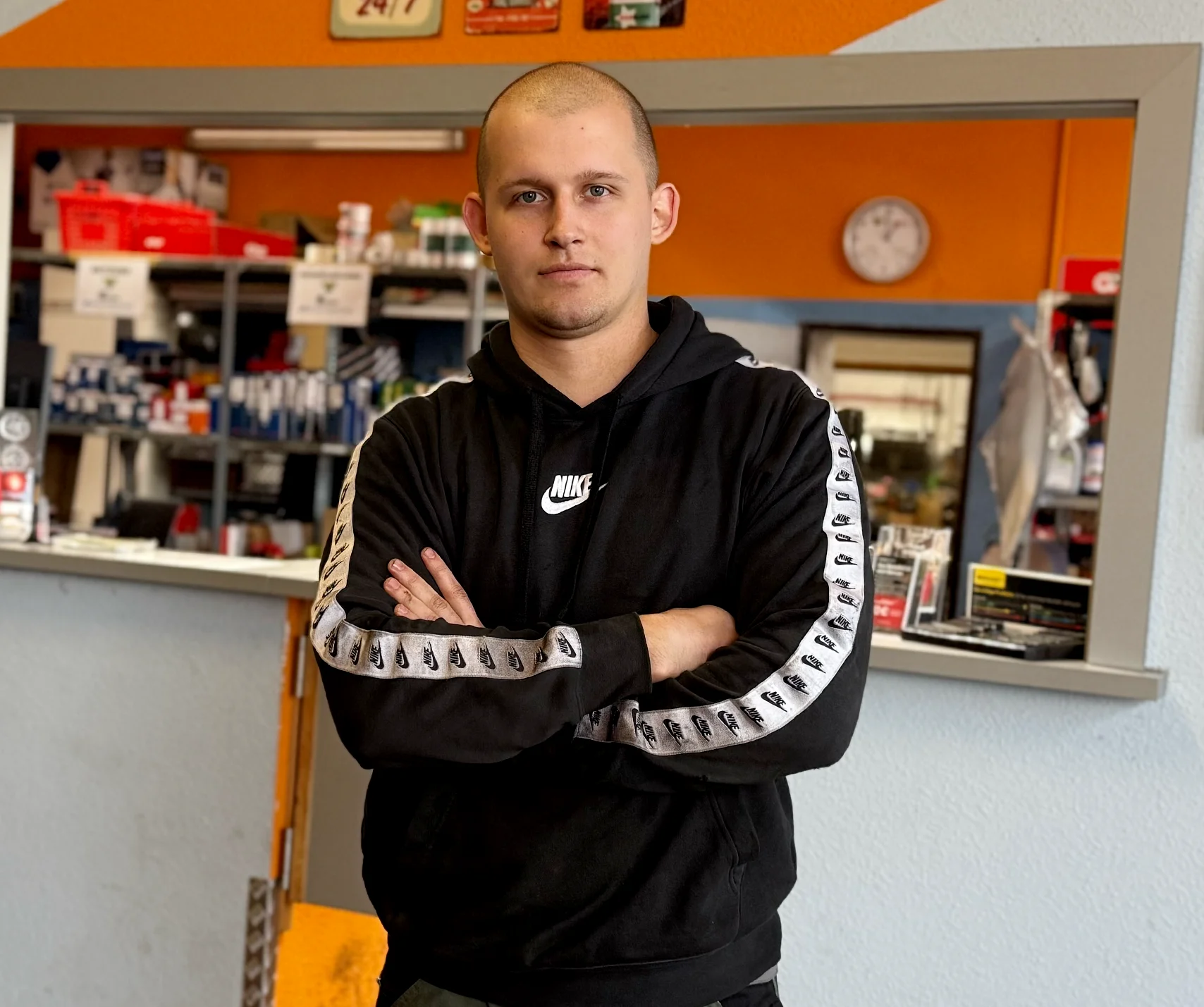 Ein Mann in Arbeitskleidung steht hin einer Werkstatt. Im Hintergrund sind Regale, Hinweisschilder und eine orangefarbene Wand zu sehen.