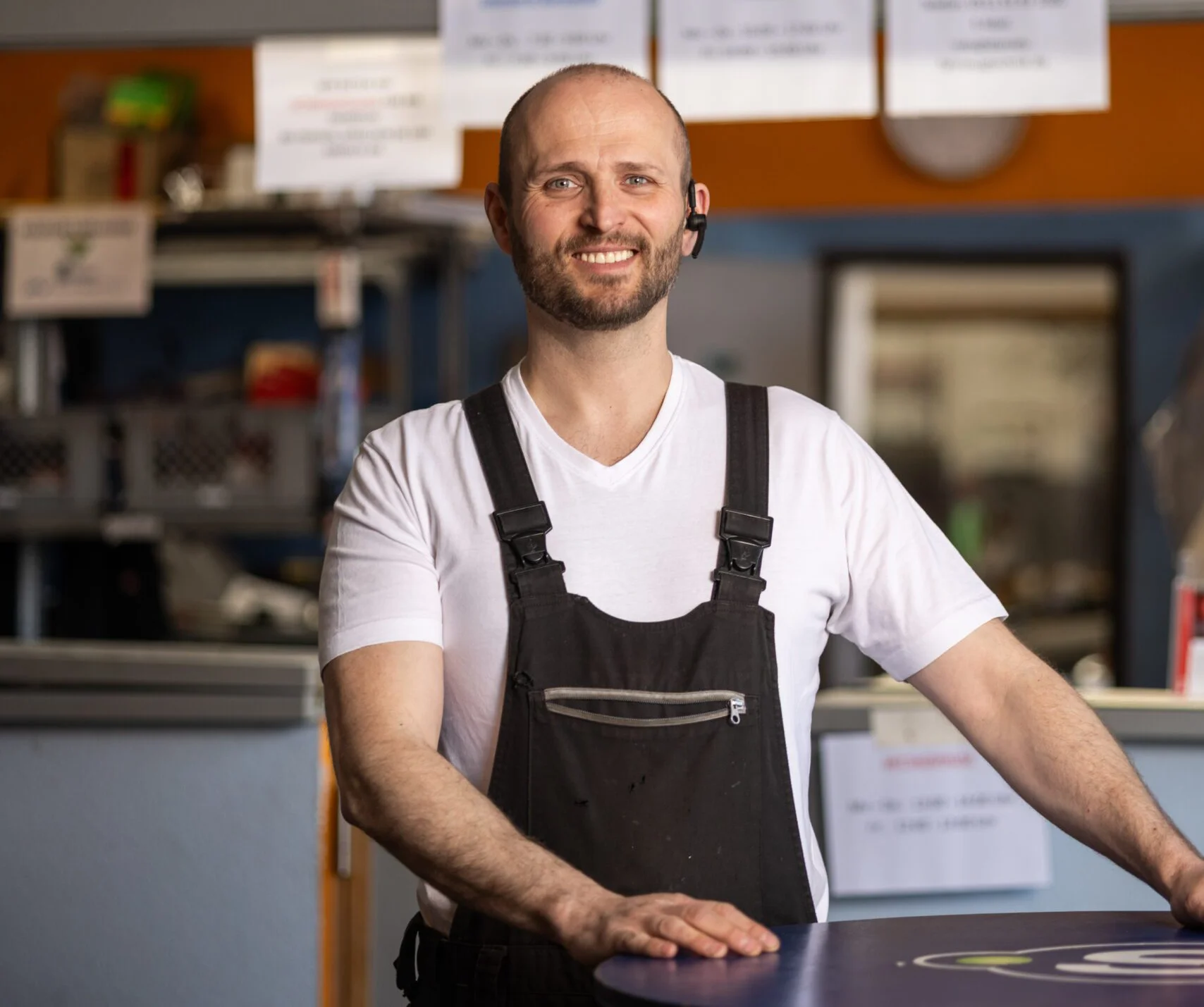 Ein Mann in Arbeitskleidung steht an einem Tresen in einer Werkstatt oder einem Geschäft, im Hintergrund sind Regale mit Werkzeugen und Hinweisschilder zu sehen.